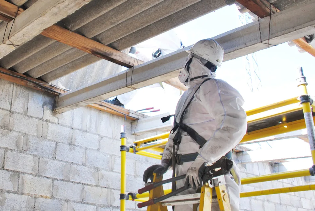 Asbestos Management and Removal Services this image shows a man in a protective suit, googles and face mask on ladders inspecting asbestos materials